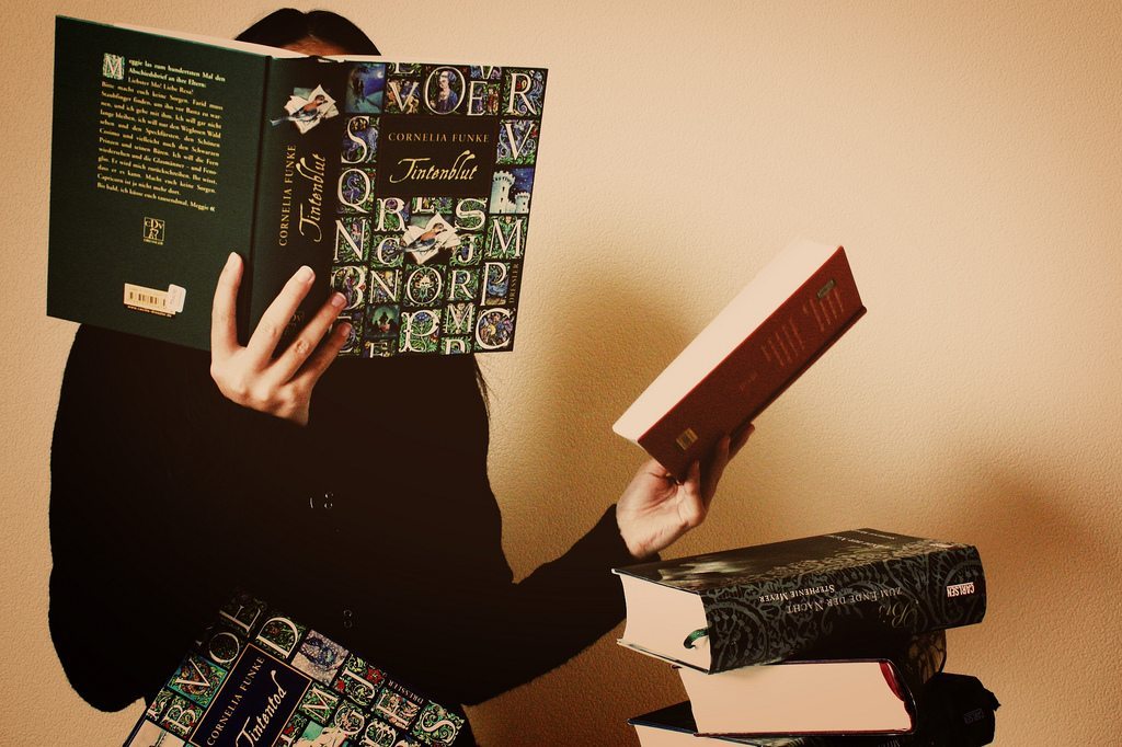 Person hidden behind a book with a large pile more to read.
