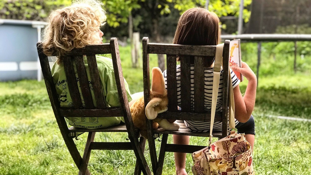 Two children facing away from camera on wooden chairs in the sunshine, one reading a book, the other looking curiously over her shoulder.