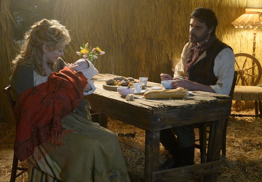 Jean Smart as Melanie Bird as a fairy-tale peasant wife, rocking a baby, and Jemaine Clements as Oliver Bird as a fairy-tale peasant husband