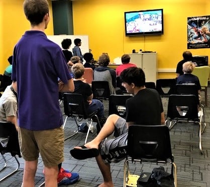 Crowd facing a large screen TV on the wall, which shows a game of Super Smash Bros in progress