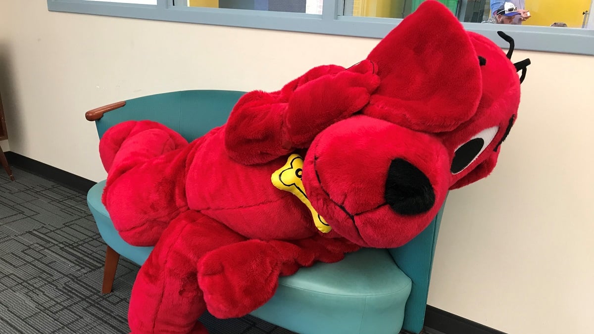 Person in a stuffed Clifford suit sprawls on a small loveseat, one ear over an eye